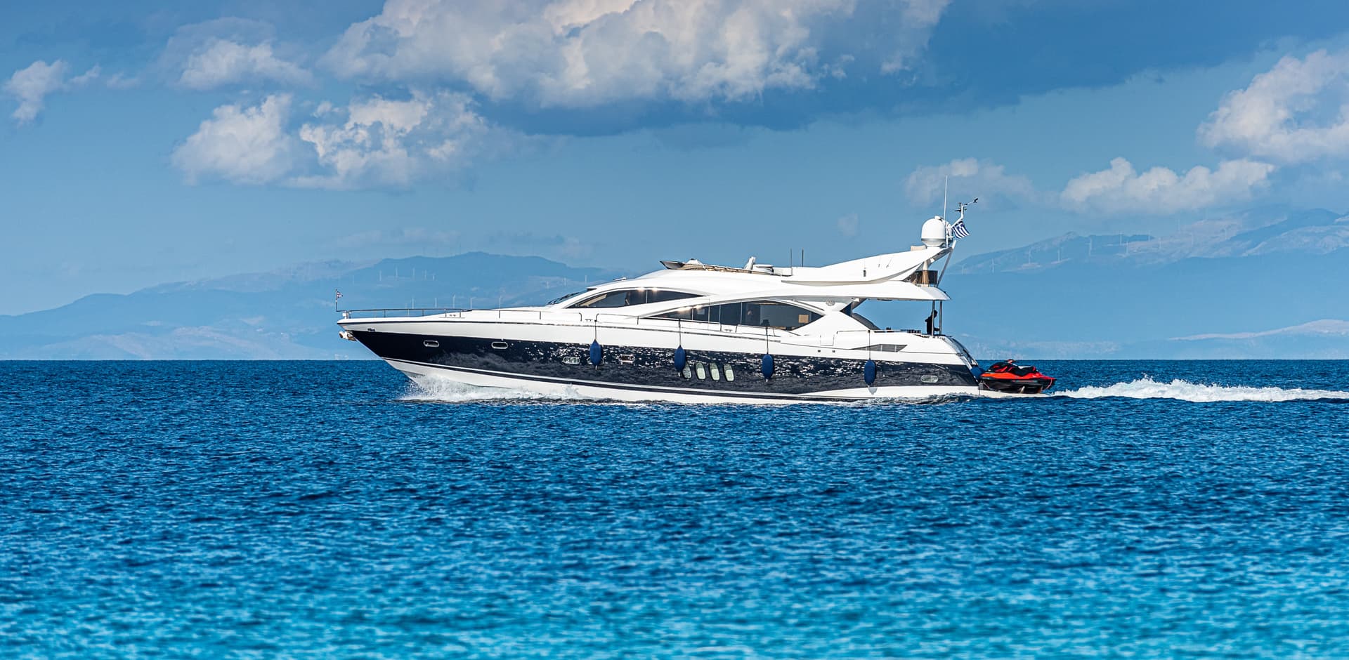 Luxury motor yacht speeding across blue water with Greek flag near hazy mountains.