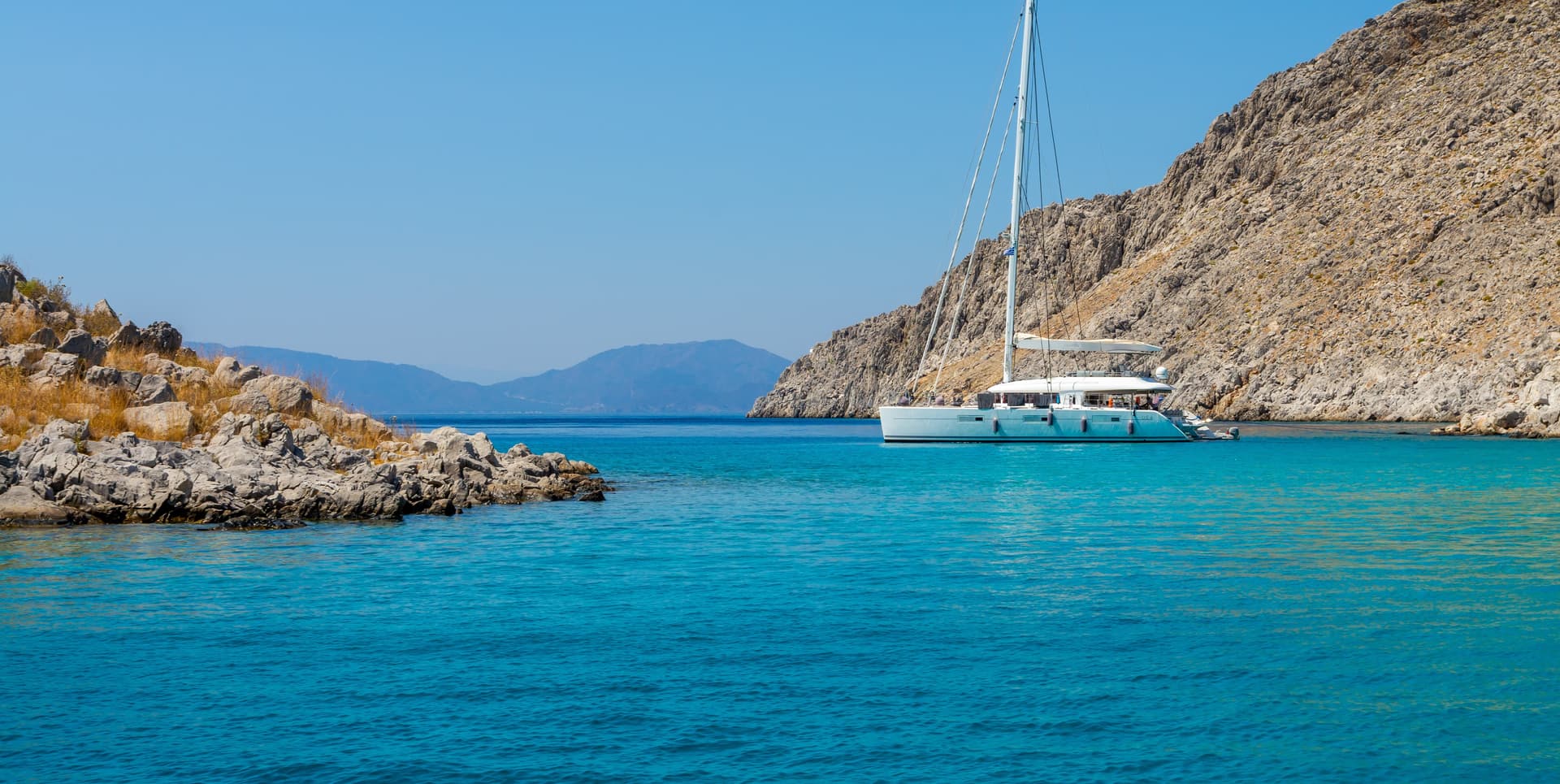 White catamaran anchored in turquoise cove between rocky, arid cliffs.