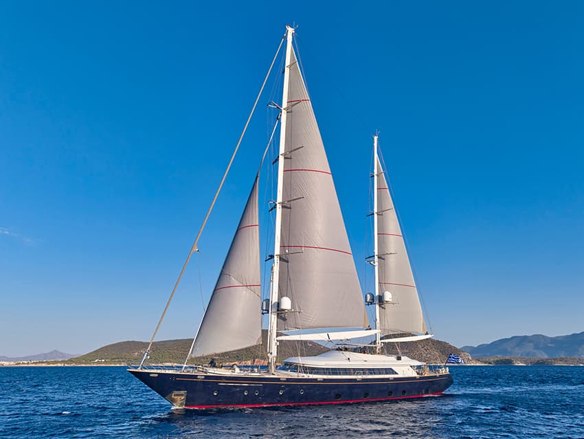 Large blue sailboat with full sails on choppy blue water near hilly coastline