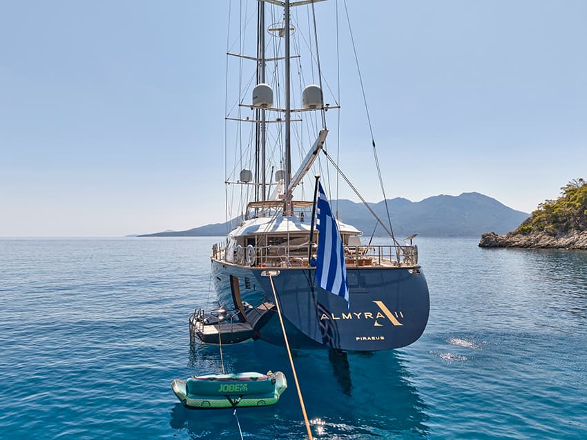 Large sailboat named ALMYRA II from Piraeus anchored in clear blue water near a mountainous coast.