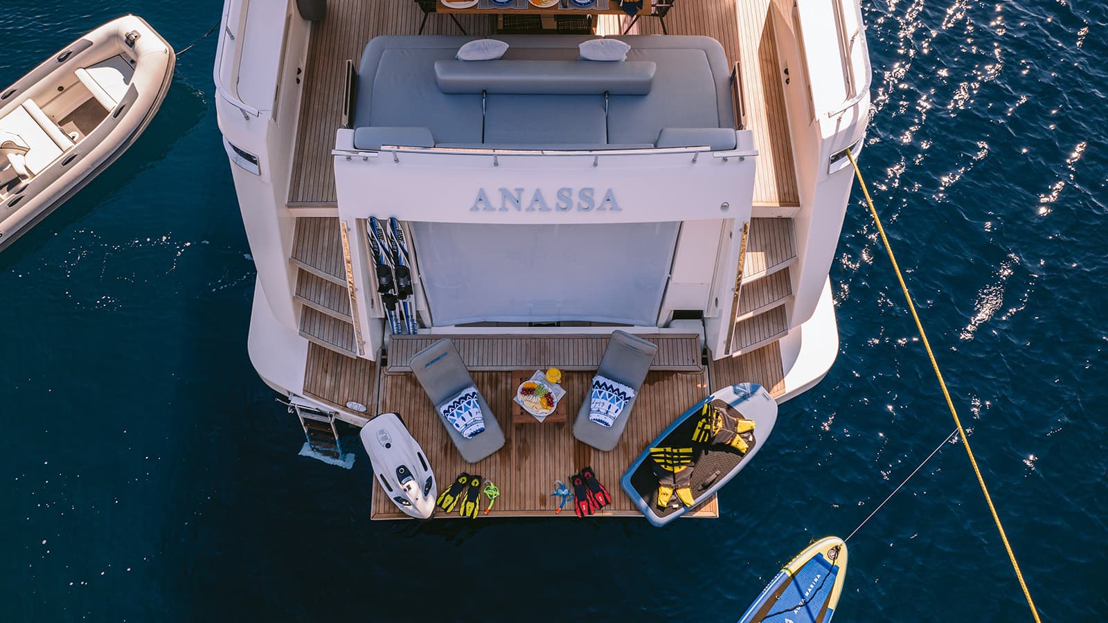Yacht "ANASSA" stern with water toys, lounge chairs, and inflatable boat on dark blue water.