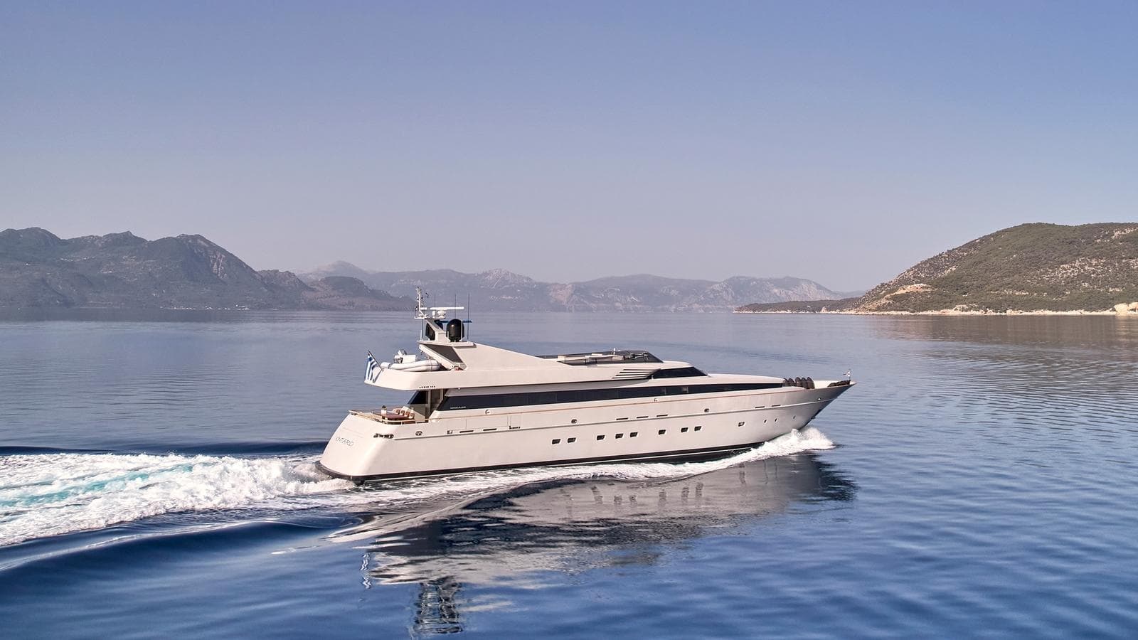 Luxury motor yacht cruising on calm blue sea near mountainous coastline under clear sky.
