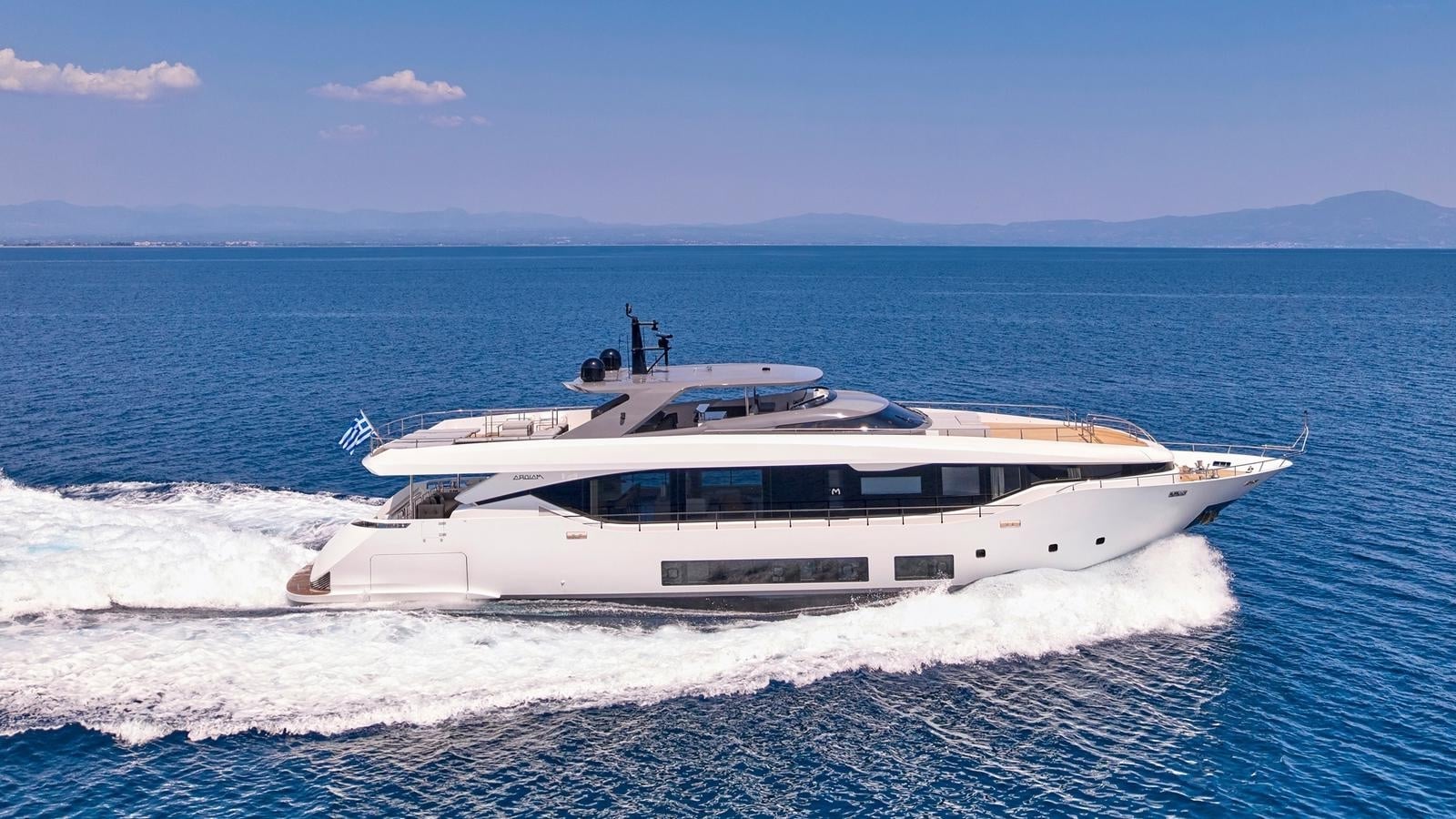 Luxury motor yacht speeding through deep blue sea with Greek flag flying near mountains.