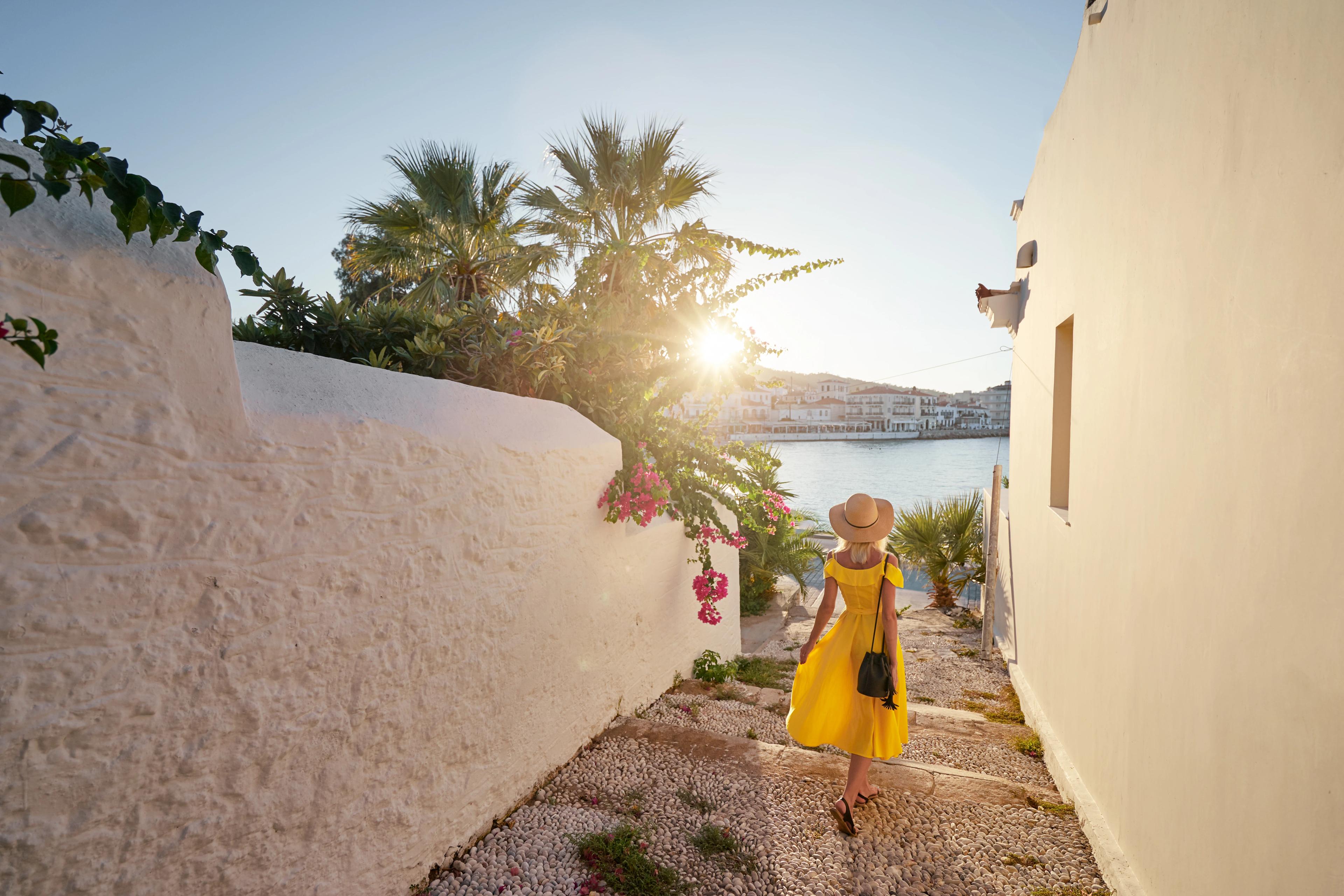 Young traveling woman in hat and yellow dress walking on old town enjoying the sea view.
