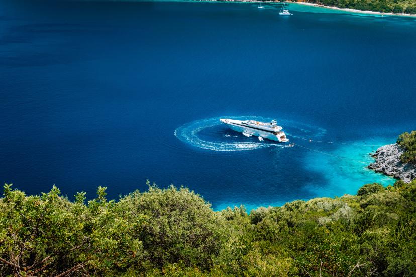 Luxury white yacht sail boat anchoring in a tranquil bay in deep blue water water, near picturesque shore of greek islands